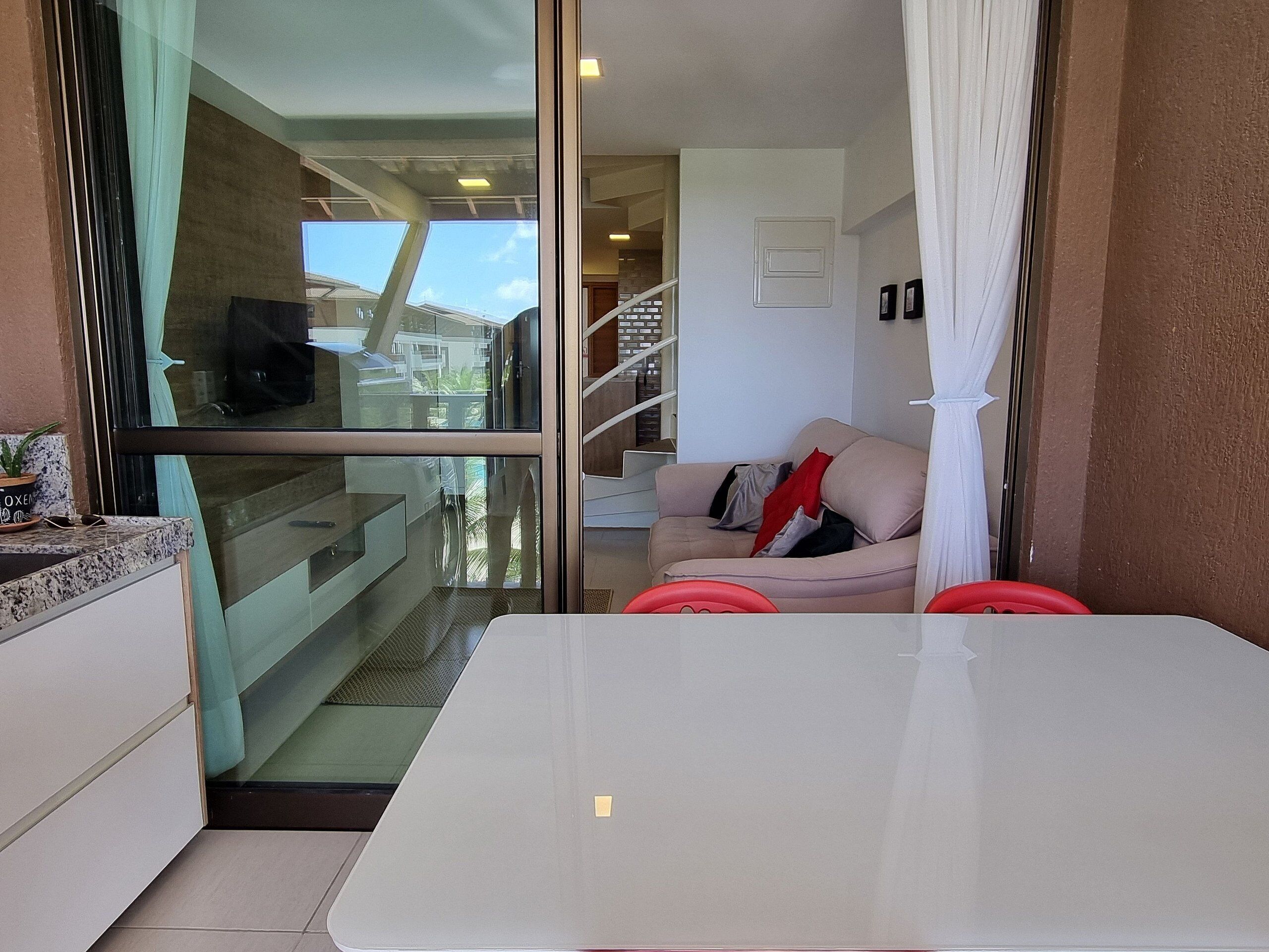 Un interior moderno con un puerta de cristal que da a un balcón, una mesa blanca y un sofá beige con cojines rojos.