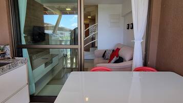 Un interior moderno con un puerta de cristal que da a un balcón, una mesa blanca y un sofá beige con cojines rojos.