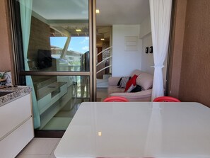 Un interior moderno con un puerta de cristal que da a un balcón, una mesa blanca y un sofá beige con cojines rojos.
