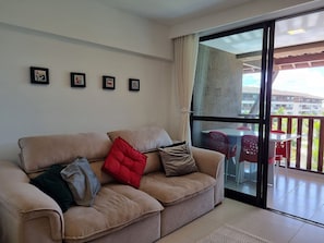 Una sala de estar con un sofá color beige, un ventanal de vidrio que da a un balcón con sillas rojas, y cuadros enmarcados en la pared.