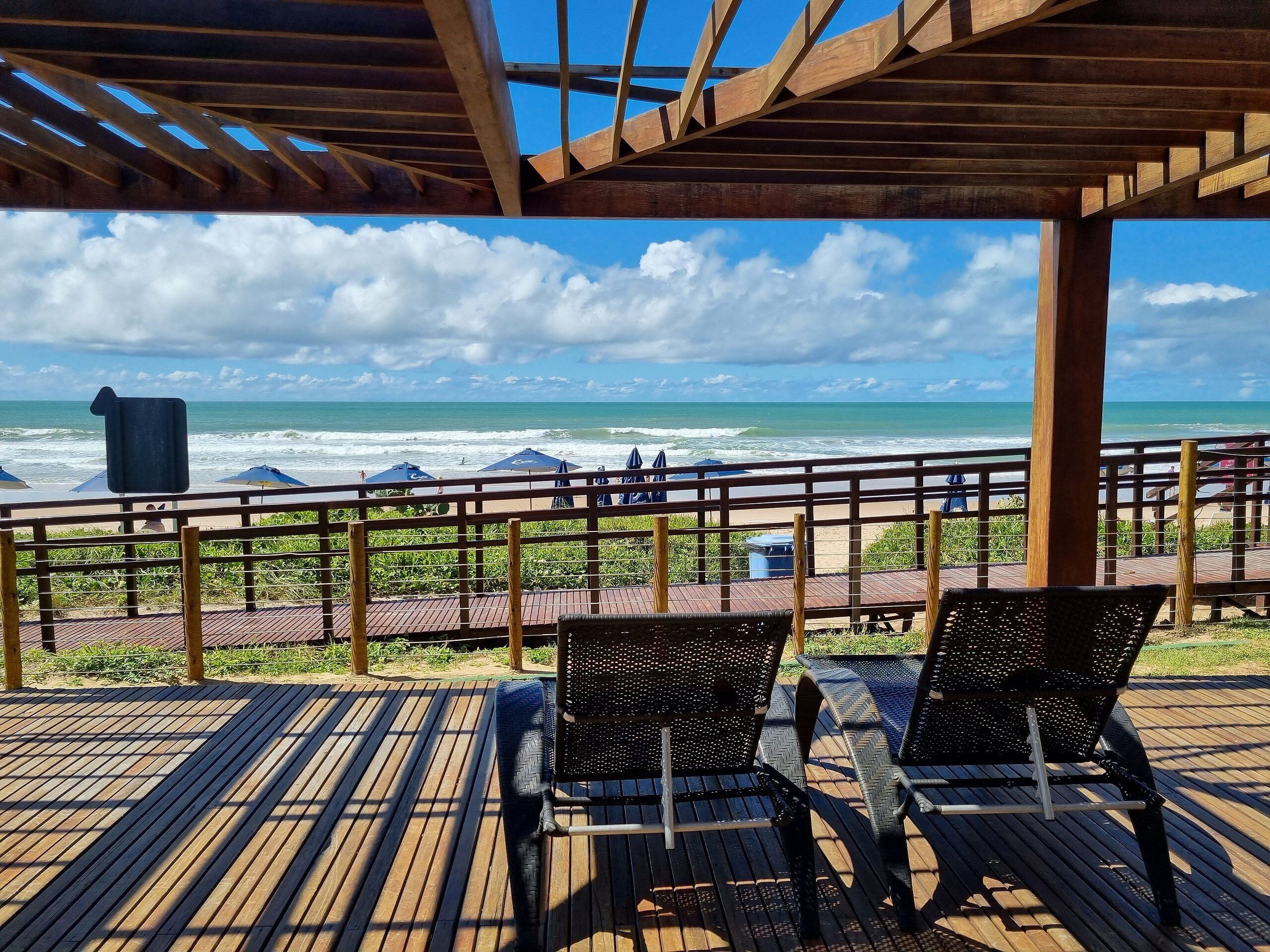 Una terraza de madera con dos sillas negras, con vista a una playa con sombrillas y el mar.