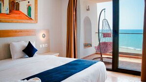 Habitación de hotel con cama, mesita de noche, luz en la pared y vista a la playa, además de una silla colgante.