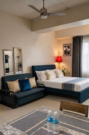 Una camera d'albergo con un letto, una poltrona, un tavolo da pranzo e bottiglie d'acqua, una ventola a soffitto e una lampada.