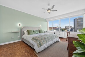 A bedroom with a bed, a ceiling fan, a window with a view of buildings, and a wooden floor. - Ocean View Apartment near V&A Waterfront (Cape Town)