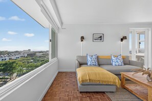 A modern living room with a sofa, two pillows, a small table, and a view of a cityscape through large windows. - Ocean View Apartment near V&A Waterfront (Cape Town)
