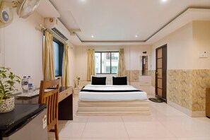 A hotel room with a bed, a desk, a chair, a potted plant, and a window with curtains.