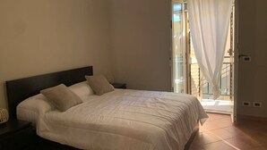 Ein Schlafzimmer mit einem Bett, einem Nachttisch und Blick auf einen Balkon durch eine offene Tür.