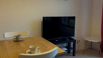 Een kamer met een houten tafel, twee witte stoelen, een televisie en twee foto's in lijsten aan de muur.