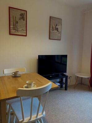 A room with a wooden table, two white chairs, a television, and two framed pictures on the wall.