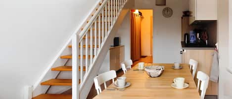 A dining area with a wooden table, white chairs, a staircase with white railings, and a kitchen area with a coffee machine.