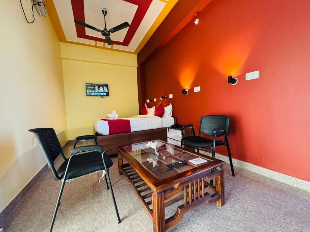 A room with a bed, a glass-top table, two chairs, a ceiling fan, and a red accent wall.