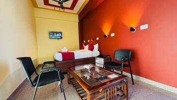 A room with a bed, a glass-top table, two chairs, a ceiling fan, and a red accent wall.