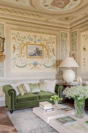 A green upholstered sofa with decorative pillows, a white table with books and a vase of flowers, and a large wall painting with foliage and birds. - Villa Maionchi: Timeless Tuscan Elegance & Luxury (Capannori)