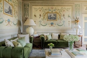 A room with green upholstered sofas, a large wall mural, and a table with books and a vase of flowers. - Villa Maionchi: Timeless Tuscan Elegance & Luxury (Capannori)