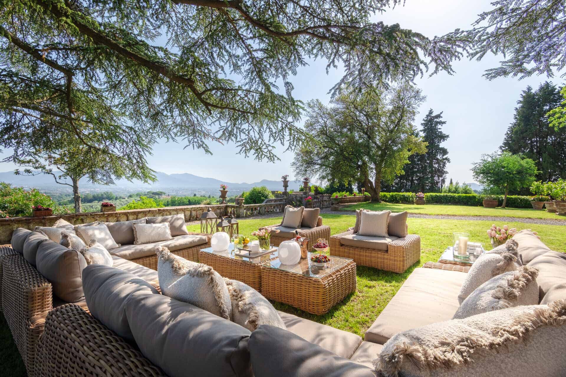 An outdoor seating area with wicker furniture and cushions, a glass table with decorative items, and a view of a landscaped garden and distant mountains.