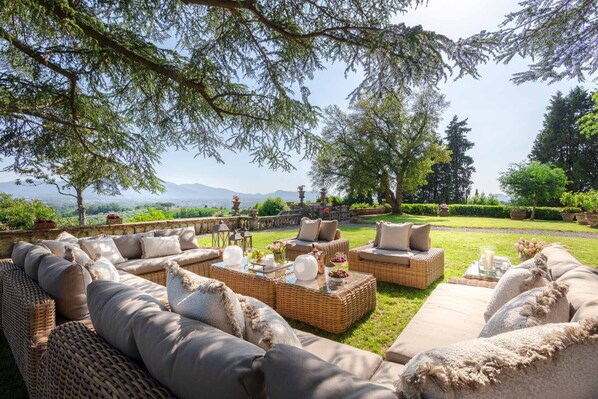 Ein Sitzbereich im Freien mit Korbmöbeln und Kissen, einem Glastisch mit Dekoration und Blick auf einen angelegten Garten sowie ferne Berge.