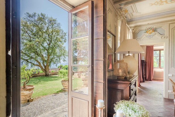 An open door leading to a room with a view of a tree and garden, a lamp, and a wooden cabinet. - Villa Maionchi: Timeless Tuscan Elegance & Luxury (Capannori)