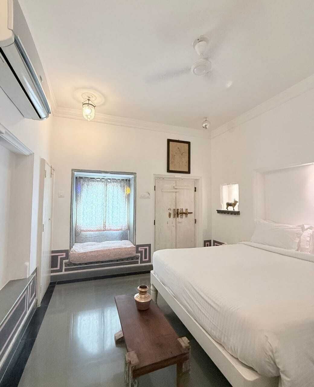 A room with a bed, a bench, a door, and a window with curtains.