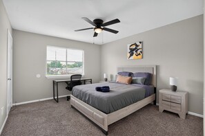 Ein Schlafzimmer mit Bett, Schreibtisch, Stuhl, Nachttisch, Lampe, Deckenventilator und einem Fenster mit Jalousien.