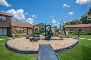 Ein Spielplatz mit Rutsche und Klettergerüst, umgeben von Kiesfläche und Rasen, mit Wohnhäusern im Hintergrund.