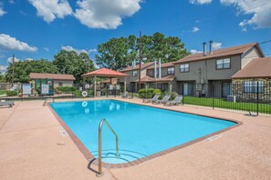 Ein Swimmingpool mit Liegestühlen und einer überdachten Sitzgelegenheit, umgeben von einem Zaun und Wohngebäuden.