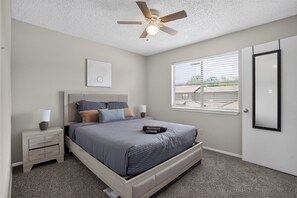 A bedroom with a bed, bedside tables, a ceiling fan, a window with blinds, and a door. - Regency Place | Modern Comfort, 2BR Home (Beaumont)