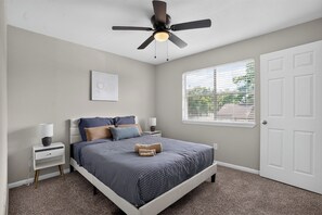 A bedroom with a bed, bedside tables, a ceiling fan, a window with blinds, and a door. - Regency Place | Fully Furnished 2BR Home (Beaumont)