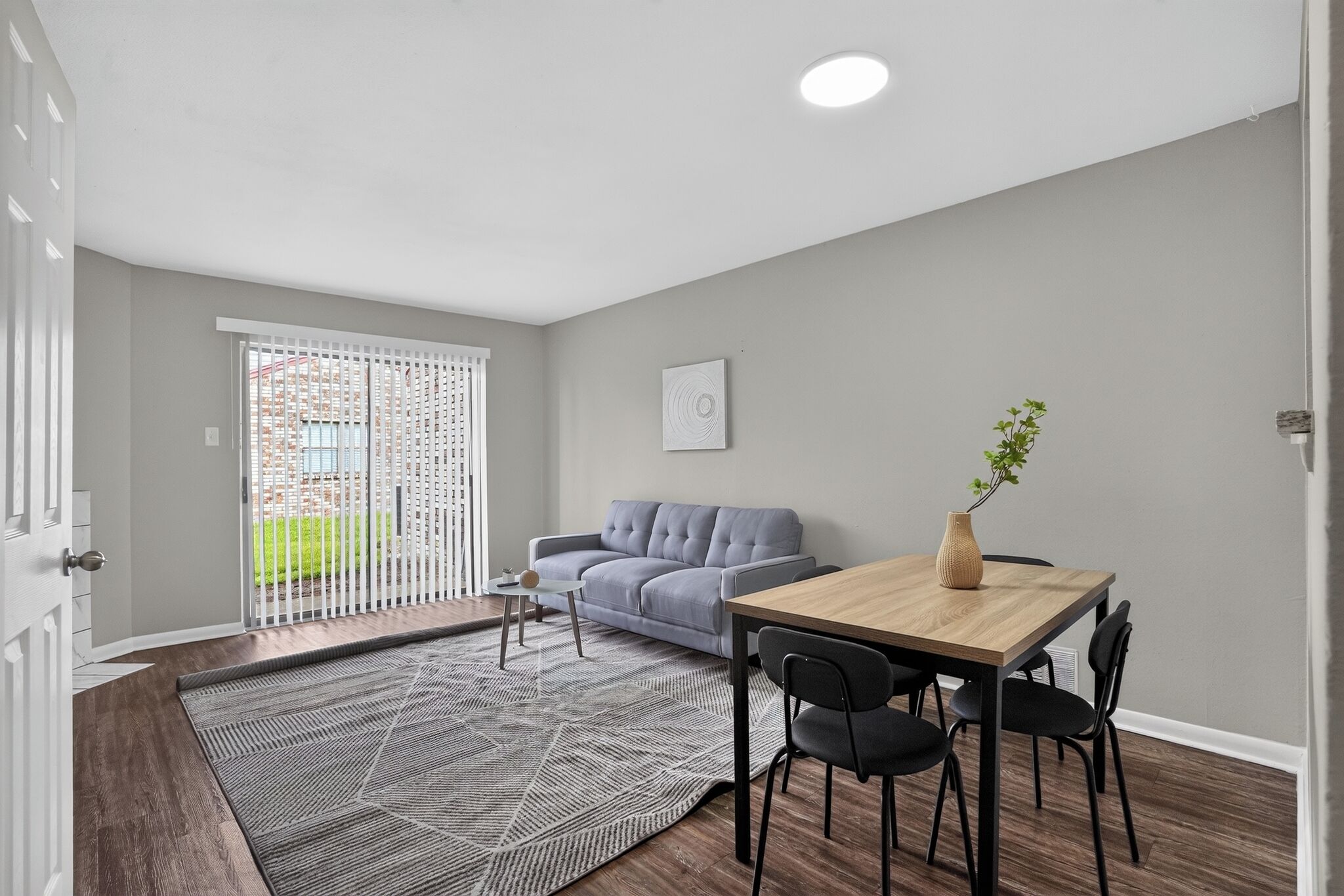 A modern living room with a grey sofa, a wooden dining table with chairs, a patterned rug, and a white door leading to a balcony with a view of greenery.