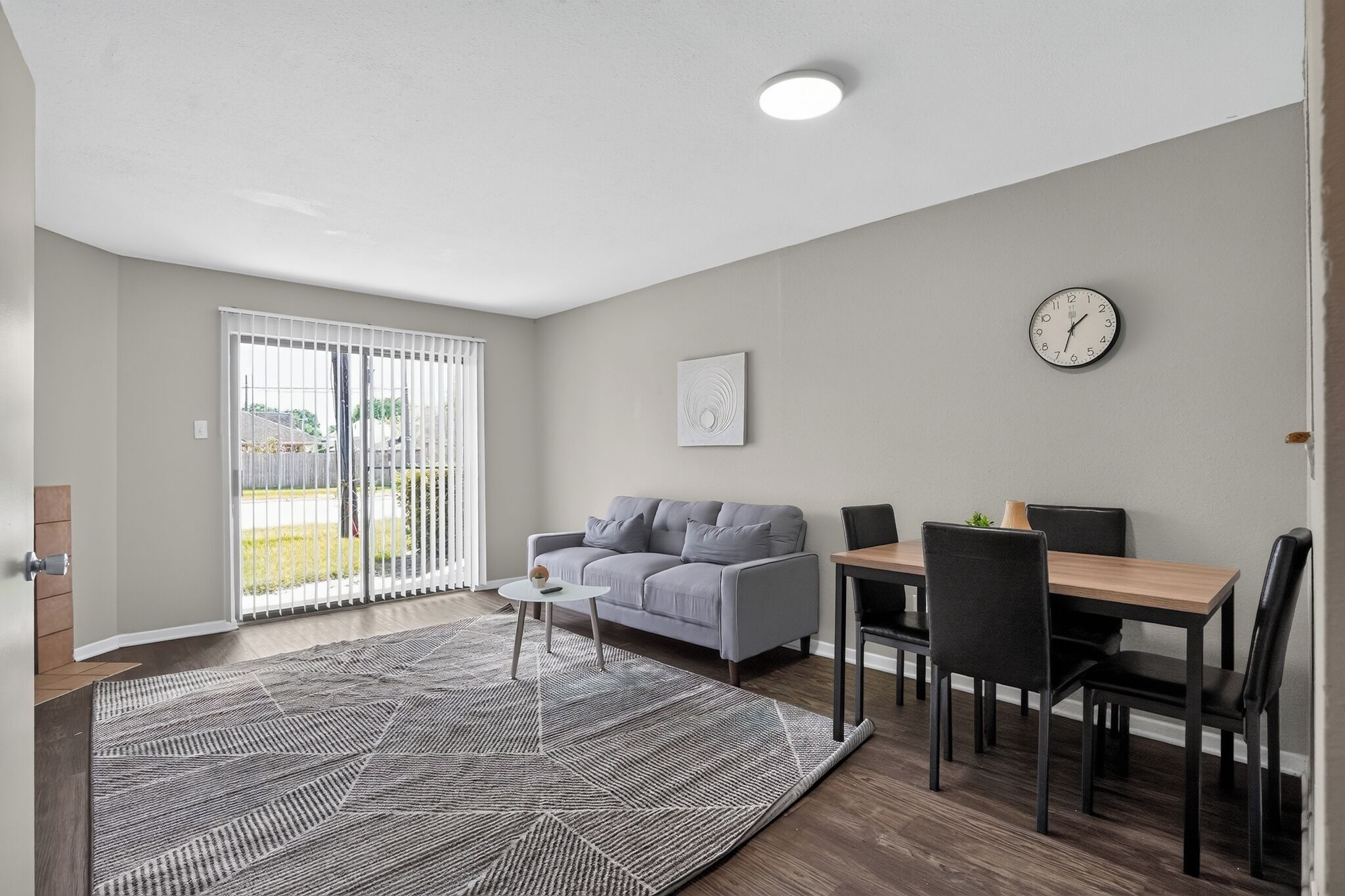 A modern living room with a grey sofa, a dining table with chairs, a patterned rug, and a ceiling light.