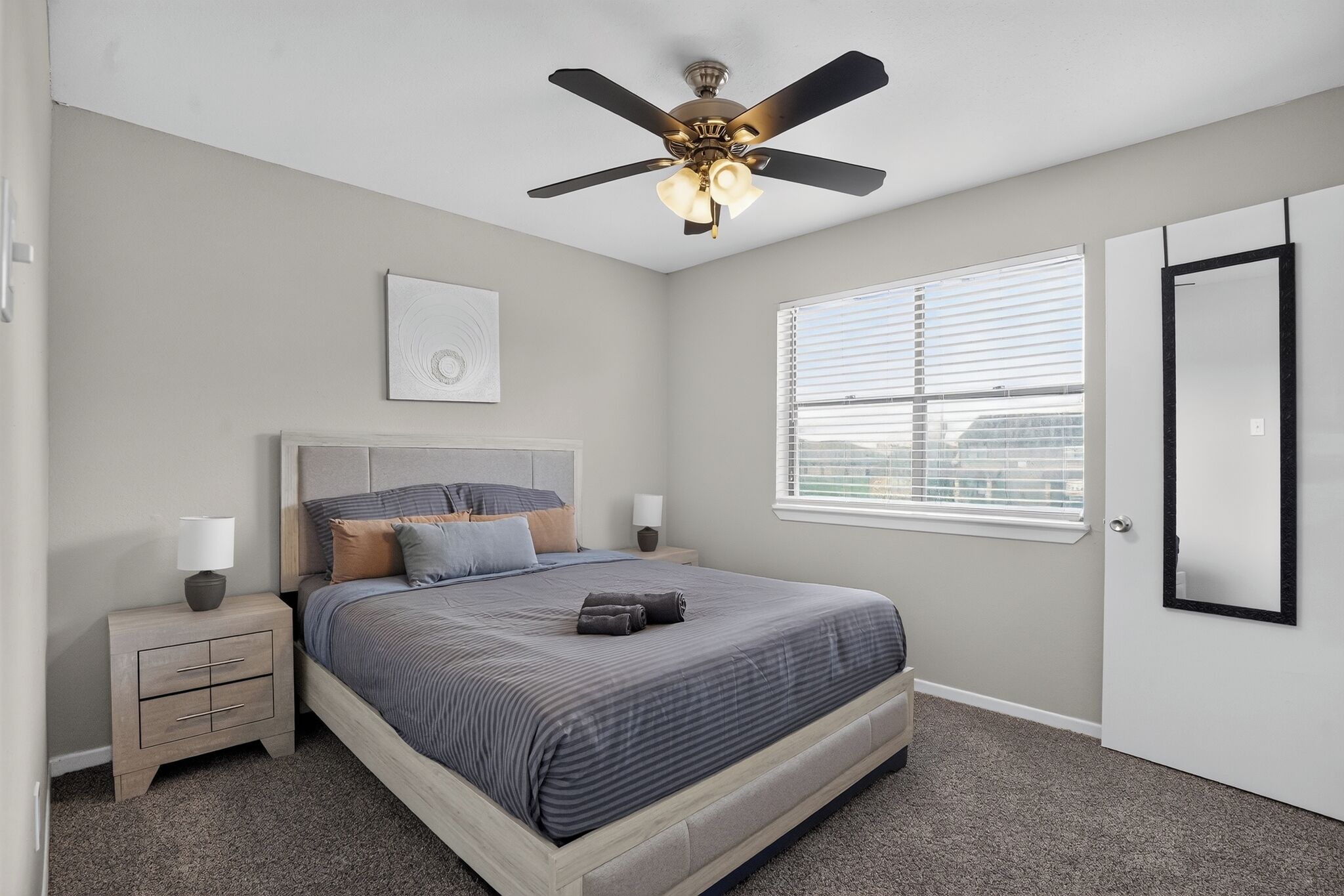 A bedroom with a bed, bedside tables, a ceiling fan, a window with blinds, and a door.