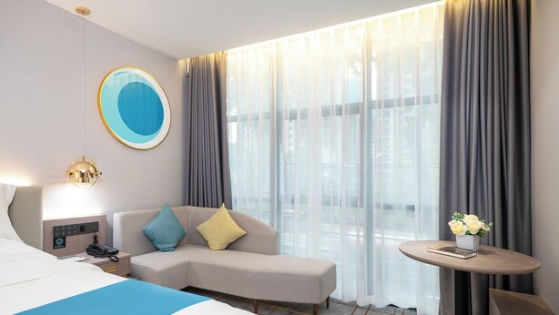 Een hotelkamer met een bed, een slaapbank, een nachtkastje met lamp, een tafeltje met een bloevas en een groot raam met gordijnen.