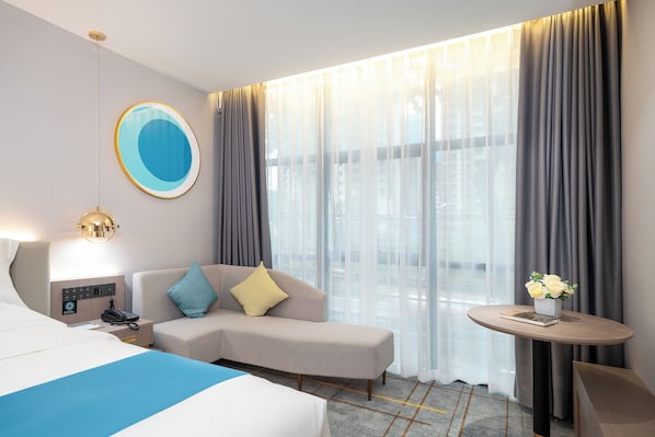 A hotel room with a bed, a sofa, a bedside table with a lamp, a small table with a vase of flowers, and a large window with curtains.