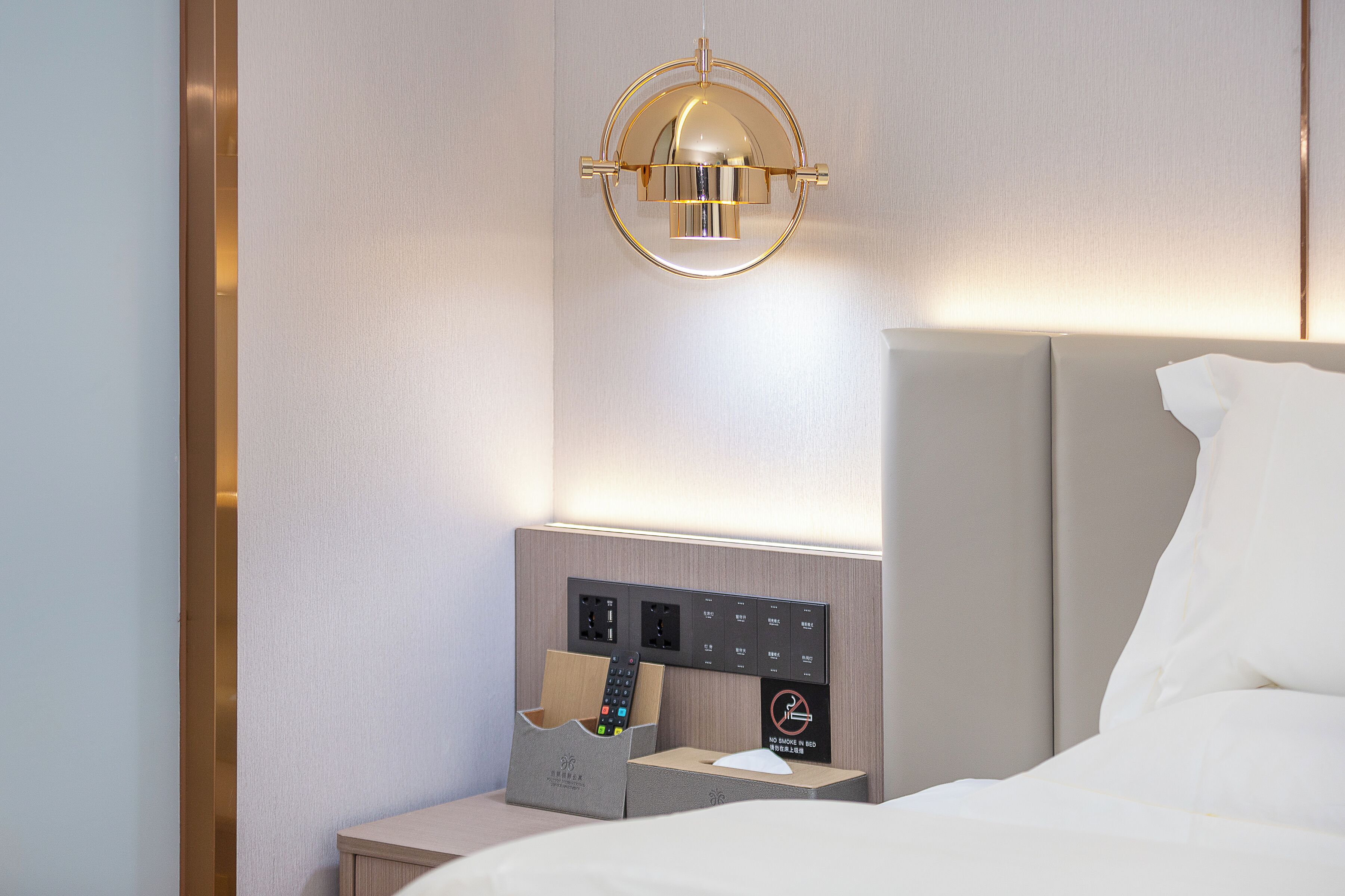 Habitación de hotel con cama, mesita de noche con lámpara y panel de control con letrero que indica 'NO FUMAR EN LA CAMA'.