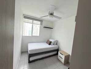 Un dormitorio pequeño y ordenado con cama, mesita de noche, ventilador de techo y ventana con vista a edificios.