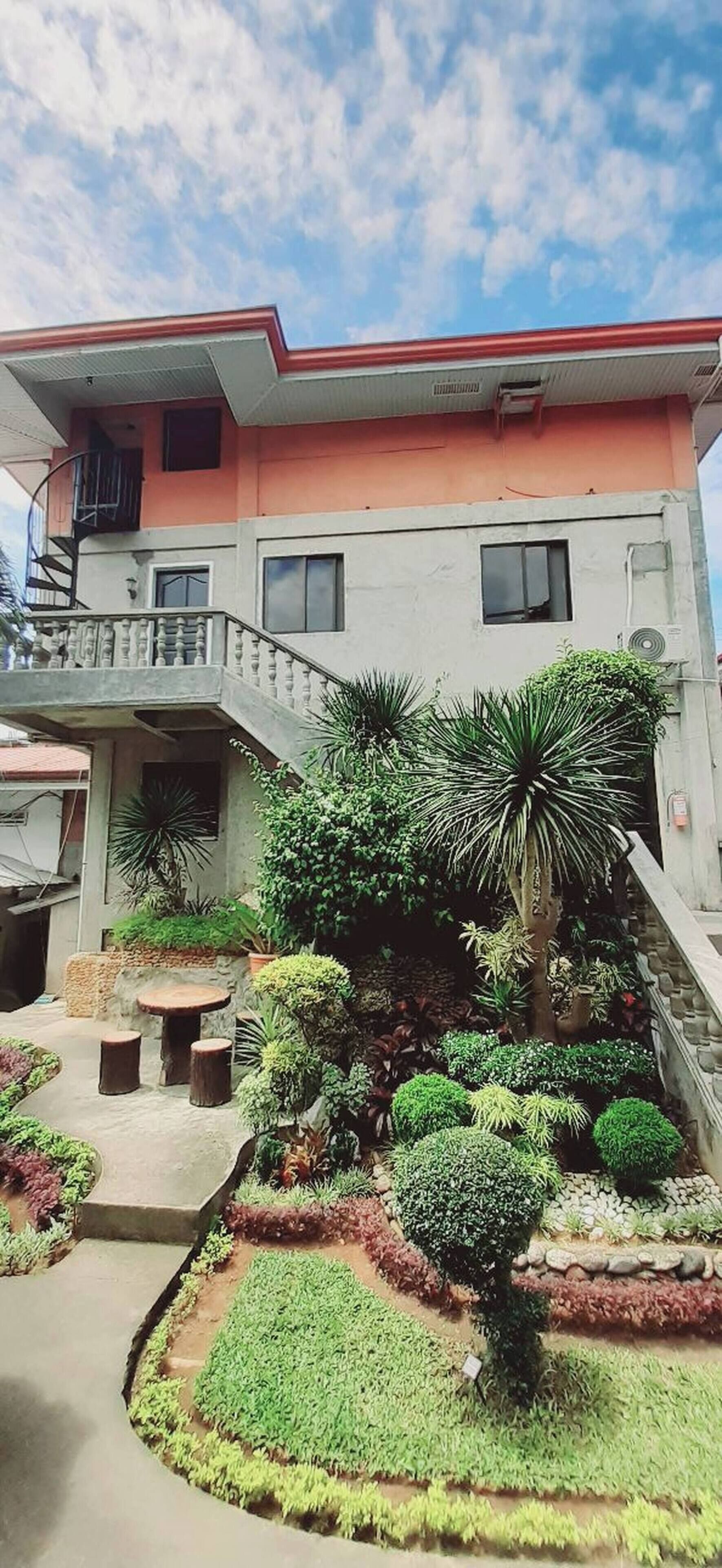 A two-story building with a balcony, a staircase leading to the upper floor, and a well-maintained garden with various plants and shrubs.
