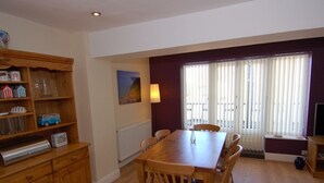 A dining area with a wooden table and chairs, a radiator, a window with blinds, and a shelf with various items. - Old Lyn Cottage (Lynmouth)