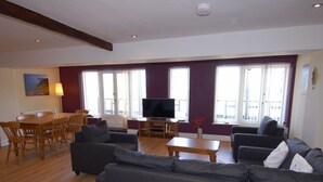A living room with a sofa, a dining table with chairs, a TV, and large windows. - Old Lyn Cottage (Lynmouth)