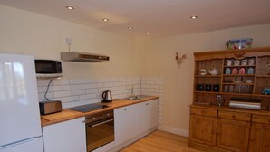 A kitchen with white cabinets, a microwave, a kettle, and wooden shelves with kitchenware. - Old Lyn Cottage (Lynmouth)