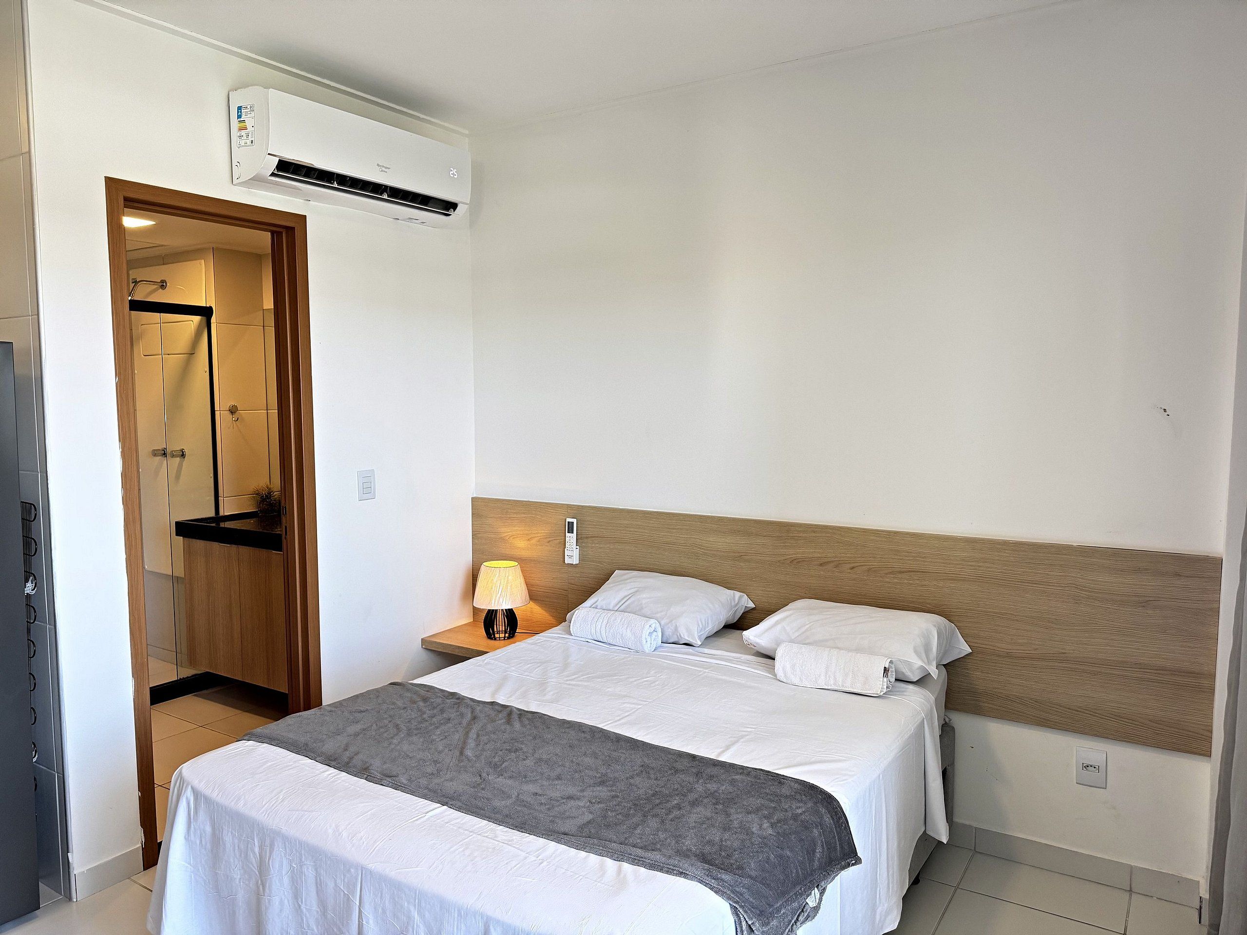 A hotel room with a bed, bedside table, lamp, and an air conditioning unit.