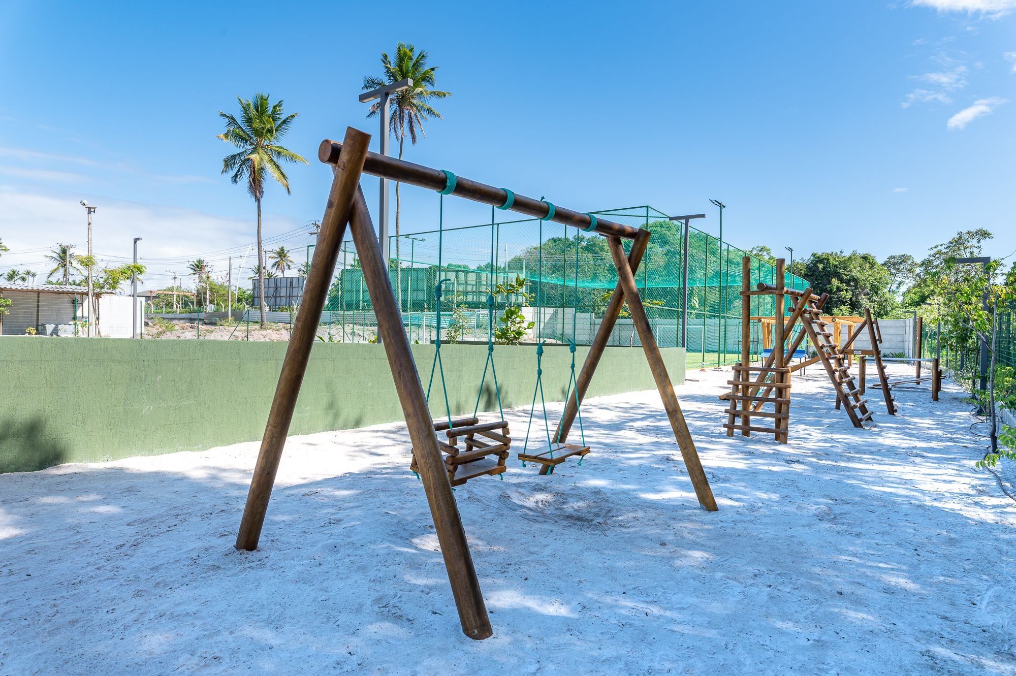 Un parque infantil con columpios y estructuras para escalar, rodeado por una cerca verde y palmeras.