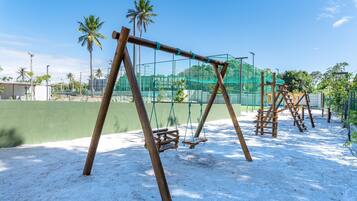 A playground with swings and climbing structures, surrounded by a green fence and palm trees.