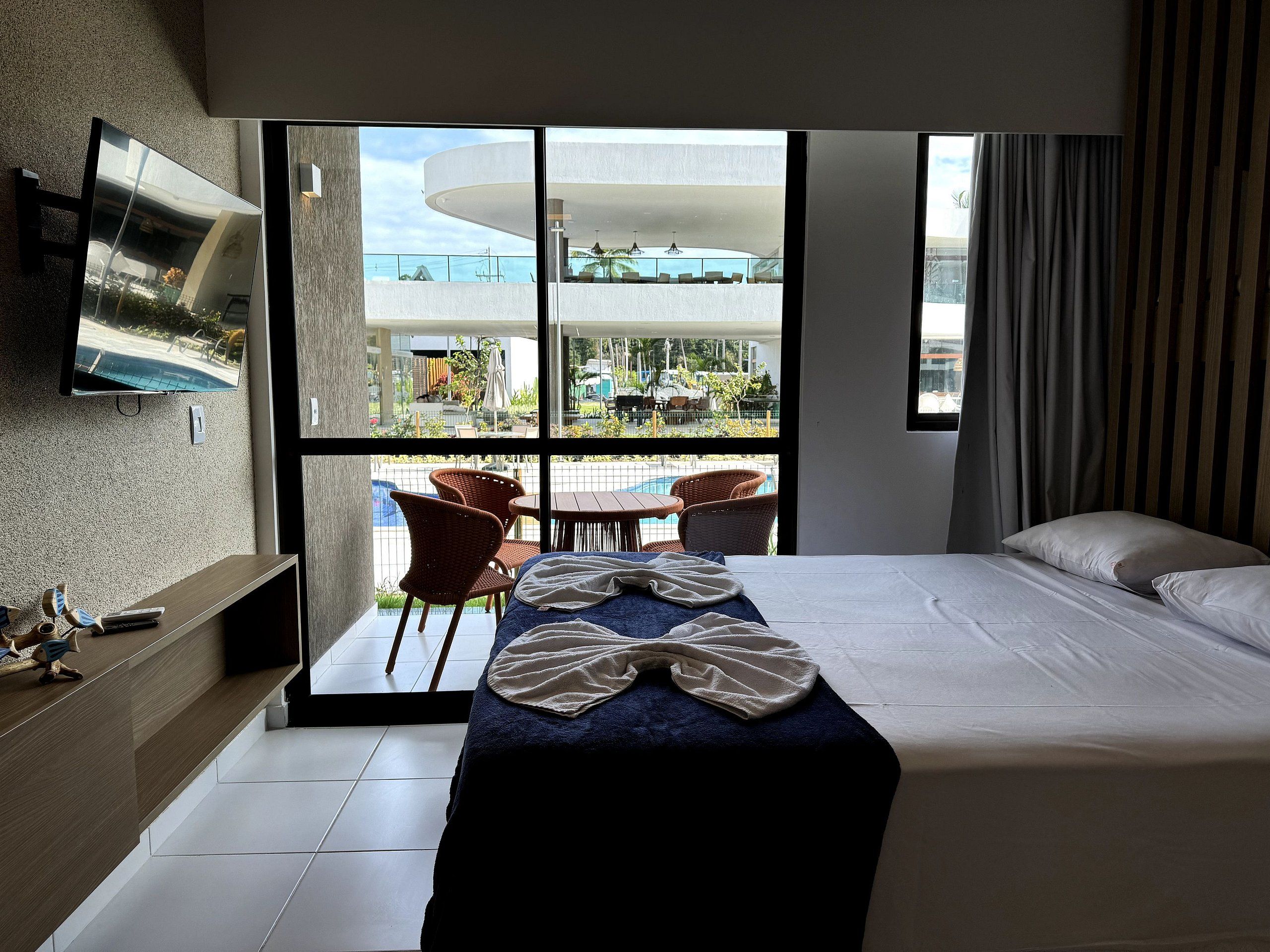 Habitación de hotel con cama, televisor montado en la pared, balcón con sillas y vista al exterior.