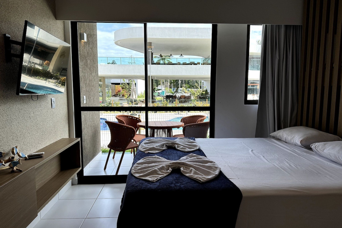 Habitación de hotel con cama, televisor montado en la pared, balcón con sillas y vista al exterior.