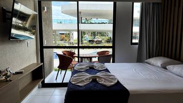 A hotel room with a bed, a TV mounted on the wall, a balcony with chairs, and a view of the outside.