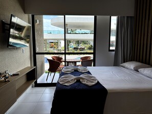 Quarto de hotel com cama, televisão na parede, varanda com cadeiras e vista para o exterior.
