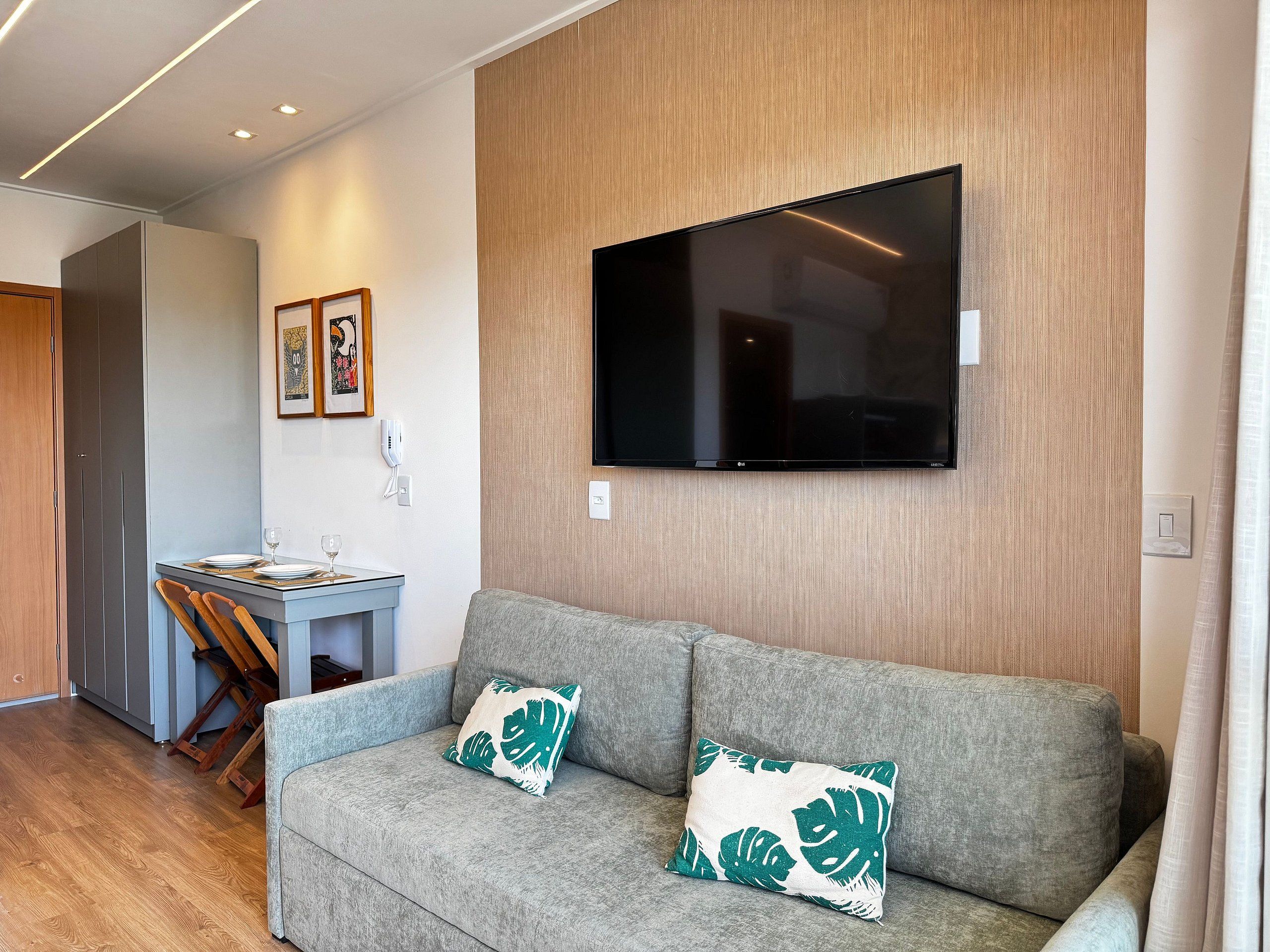 Uma sala de estar moderna com um sofá cinzento, duas almofadas decorativas, uma TV de tela plana na parede, uma mesinha com tampo de vidro e piso de madeira.