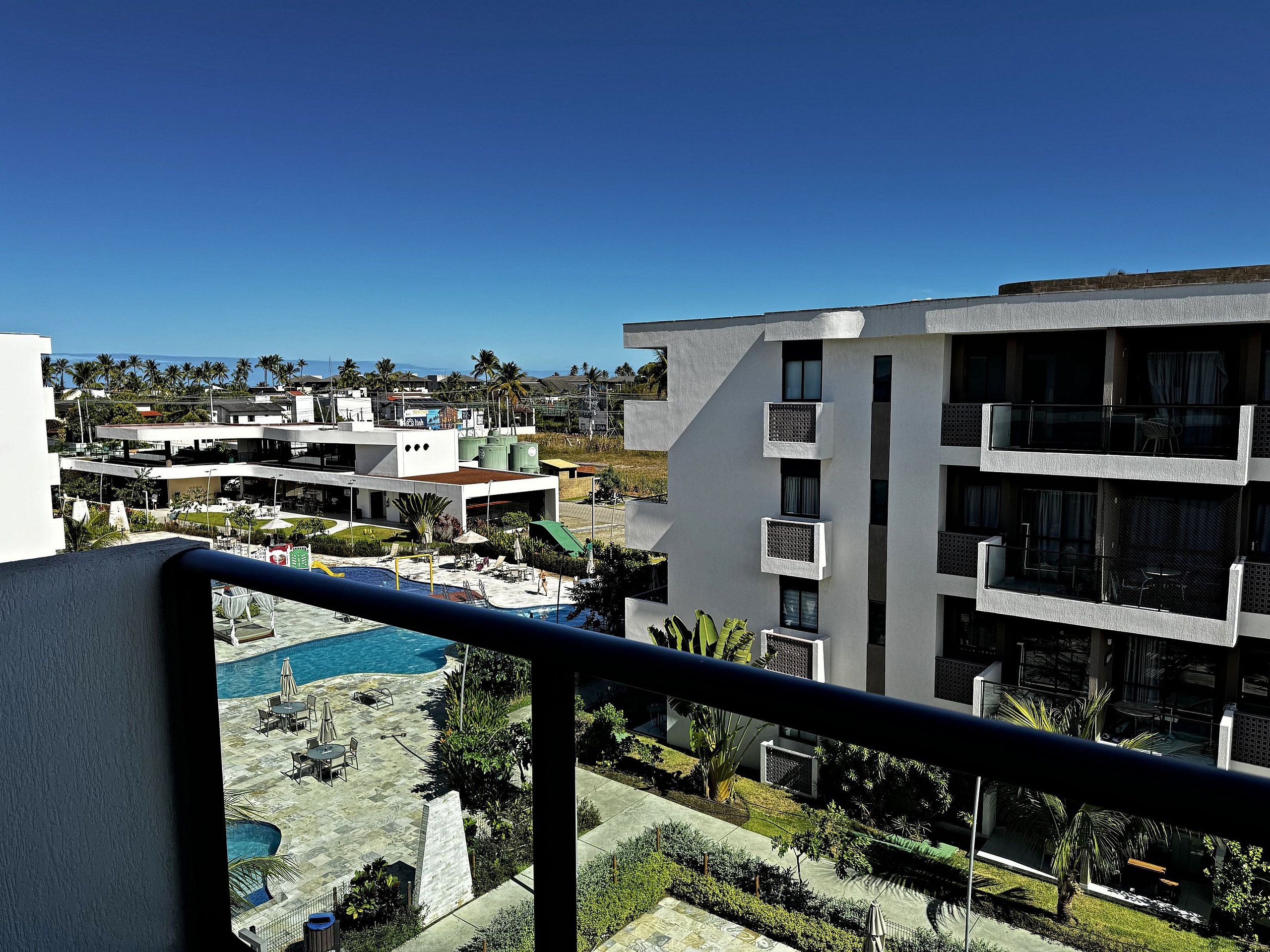 A multi-story building with balconies, a swimming pool, and a view of palm trees and other buildings.