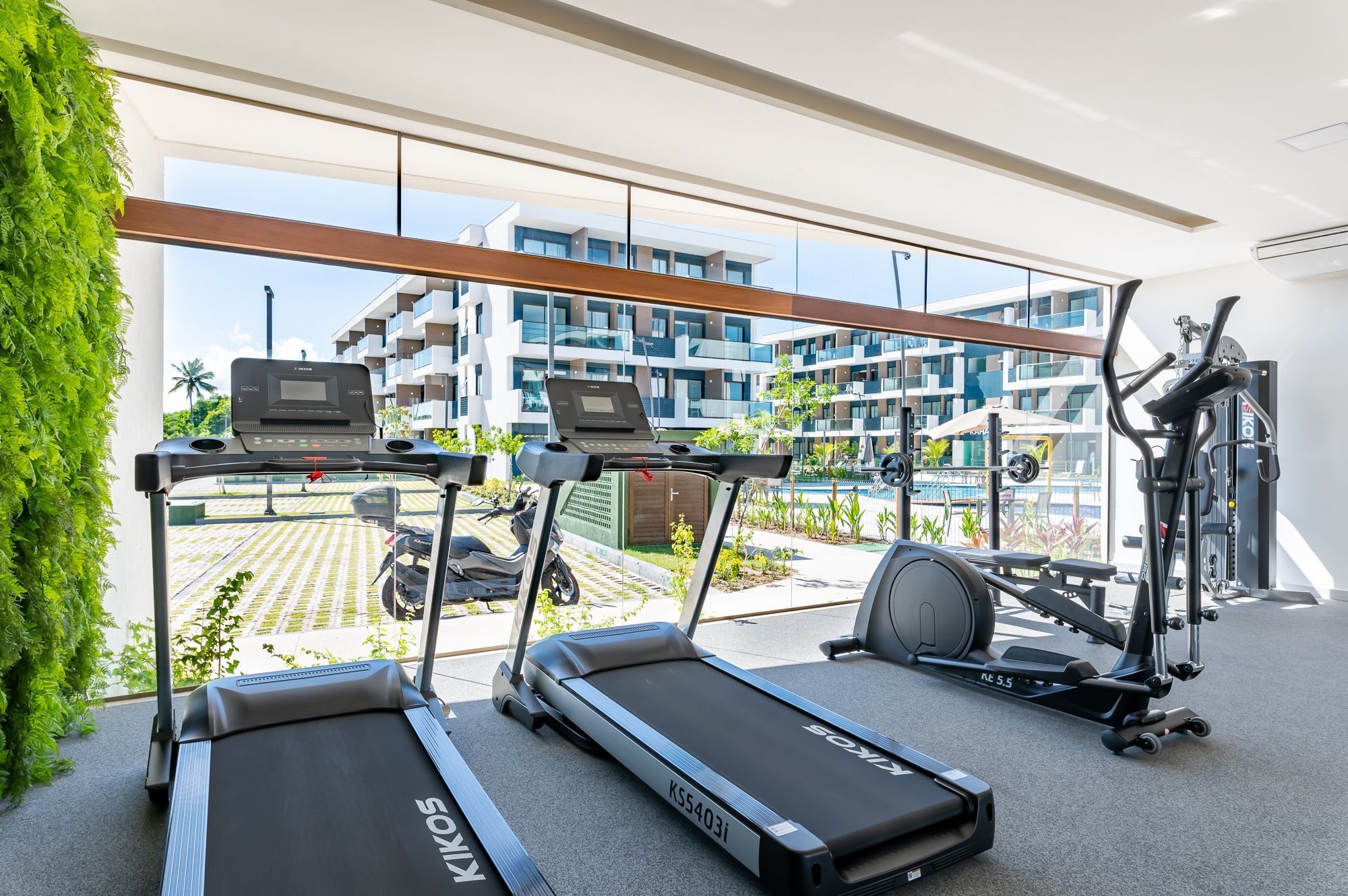 A gym with treadmills, a stationary bike, and a rowing machine, with a view of a residential building and a pool.