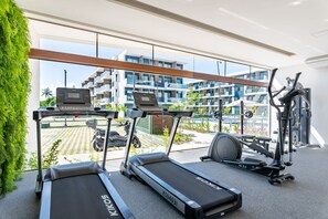 Uma academia com esteiras, bicicleta ergométrica e remo, com vista para um prédio residencial e uma piscina.