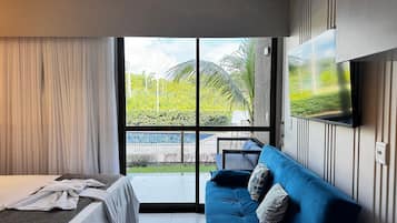 A modern hotel room with a bed, a blue sofa, a TV mounted on the wall, and a large sliding glass door leading to an outdoor area.